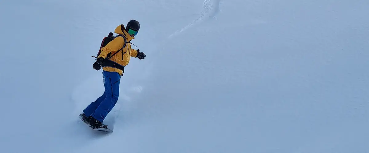 A snowboarder with a yellow jacket and blue pants going down the mountain
